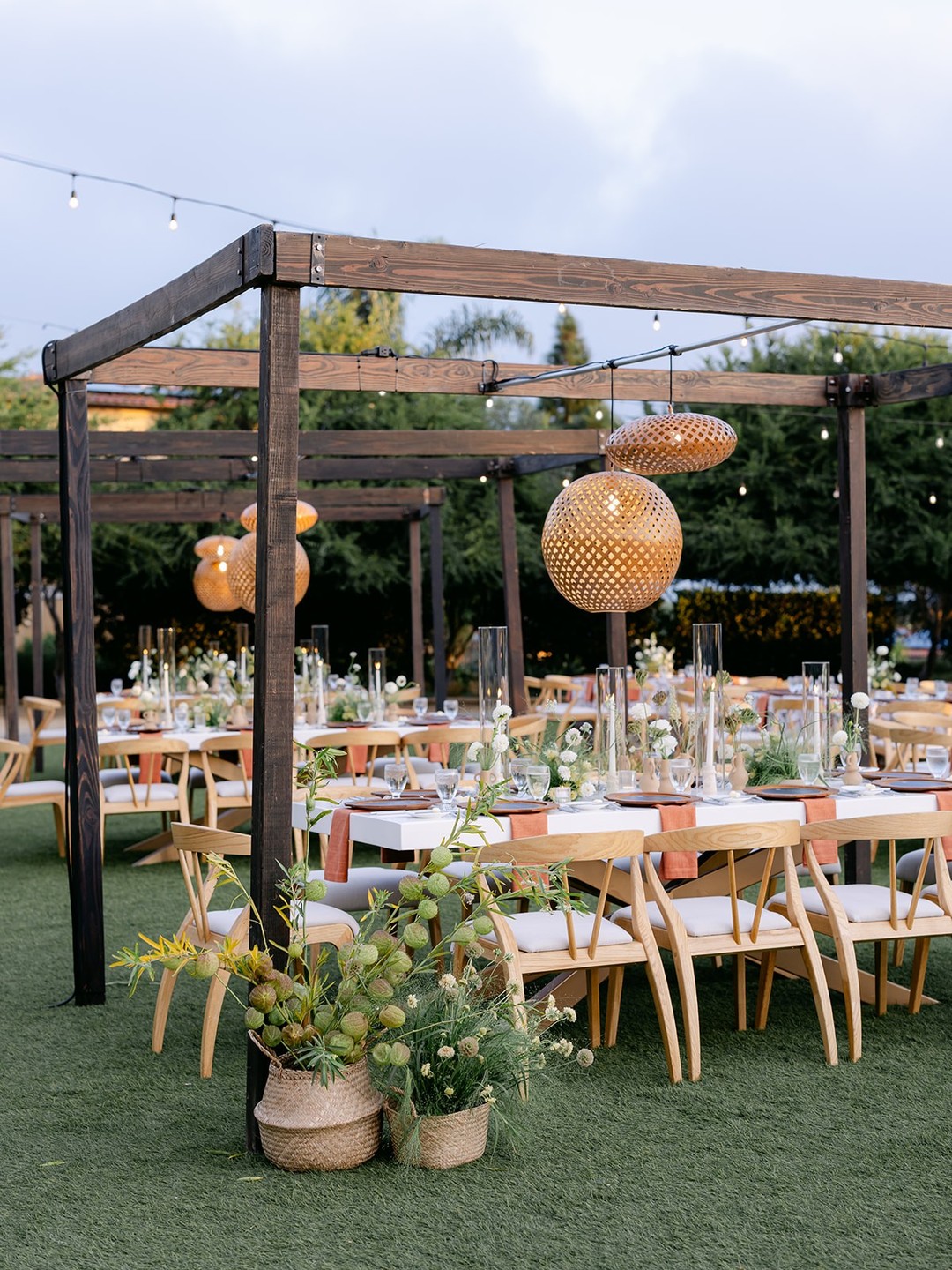 Outdoor dining area with wooden tables set with plates, glasses and flowers, wicker pendant lights, and wooden chairs on green grass.