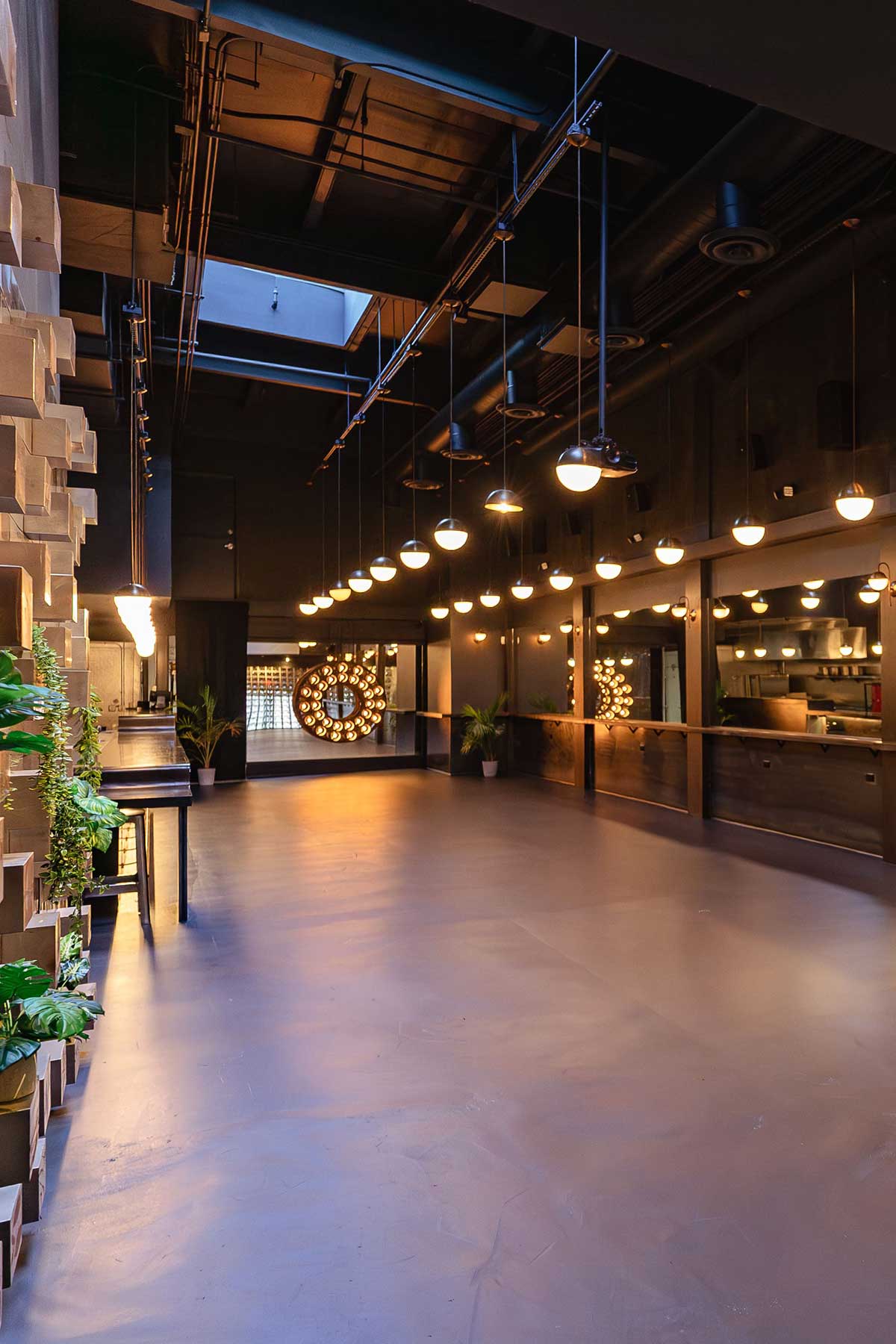 Spacious modern interior with hanging round lights, wooden wall panels, and indoor plants along the left wall.