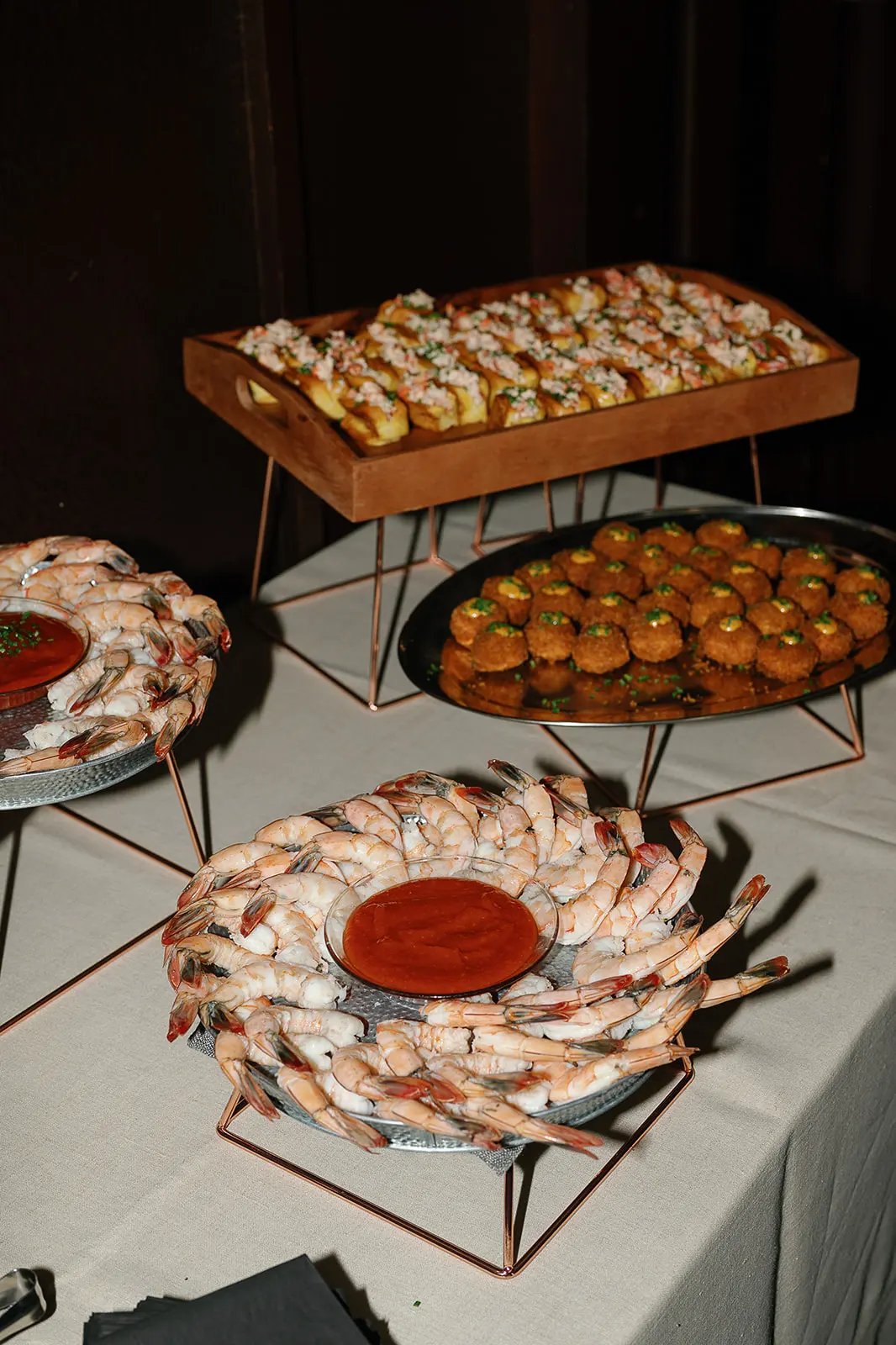 Catering setup with platters of shrimp cocktail with cocktail sauce, crab-topped bread, and fried appetizers on a table with white tablecloth.