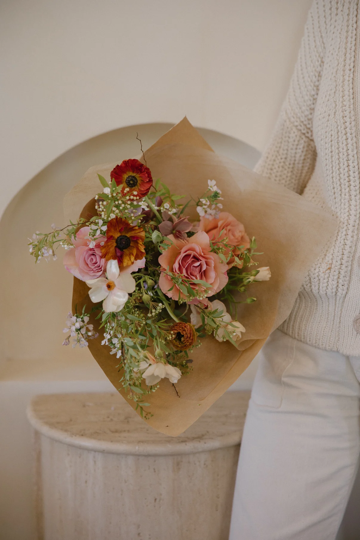 Person wearing a cream knitted sweater holding a bouquet of mixed flowers wrapped in brown paper.