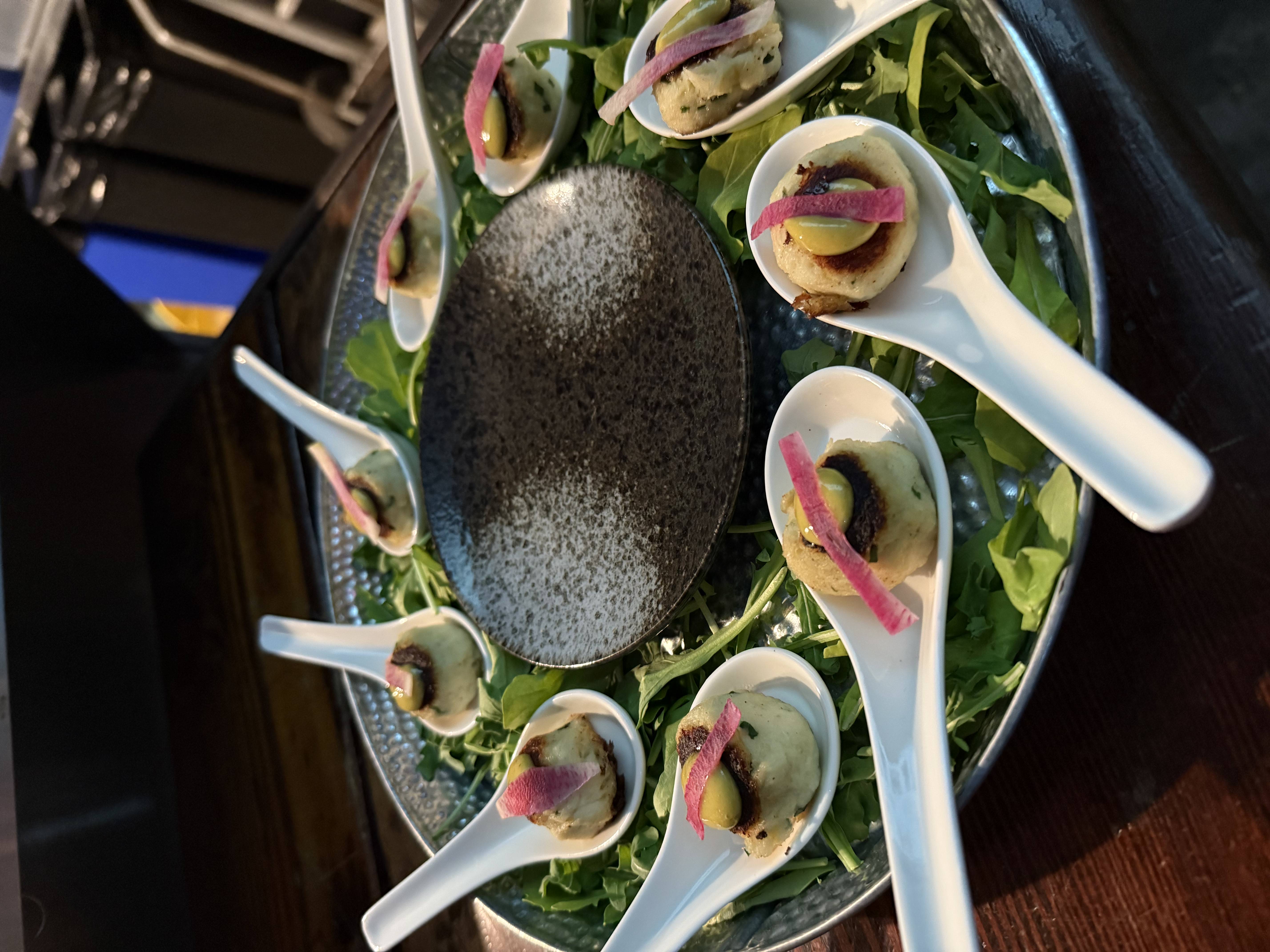 Appetizer spoons each holding a small round crab cake topped with a green dollop and a thin slice of pink garnish, arranged on a bed of greens around a textured black plate.