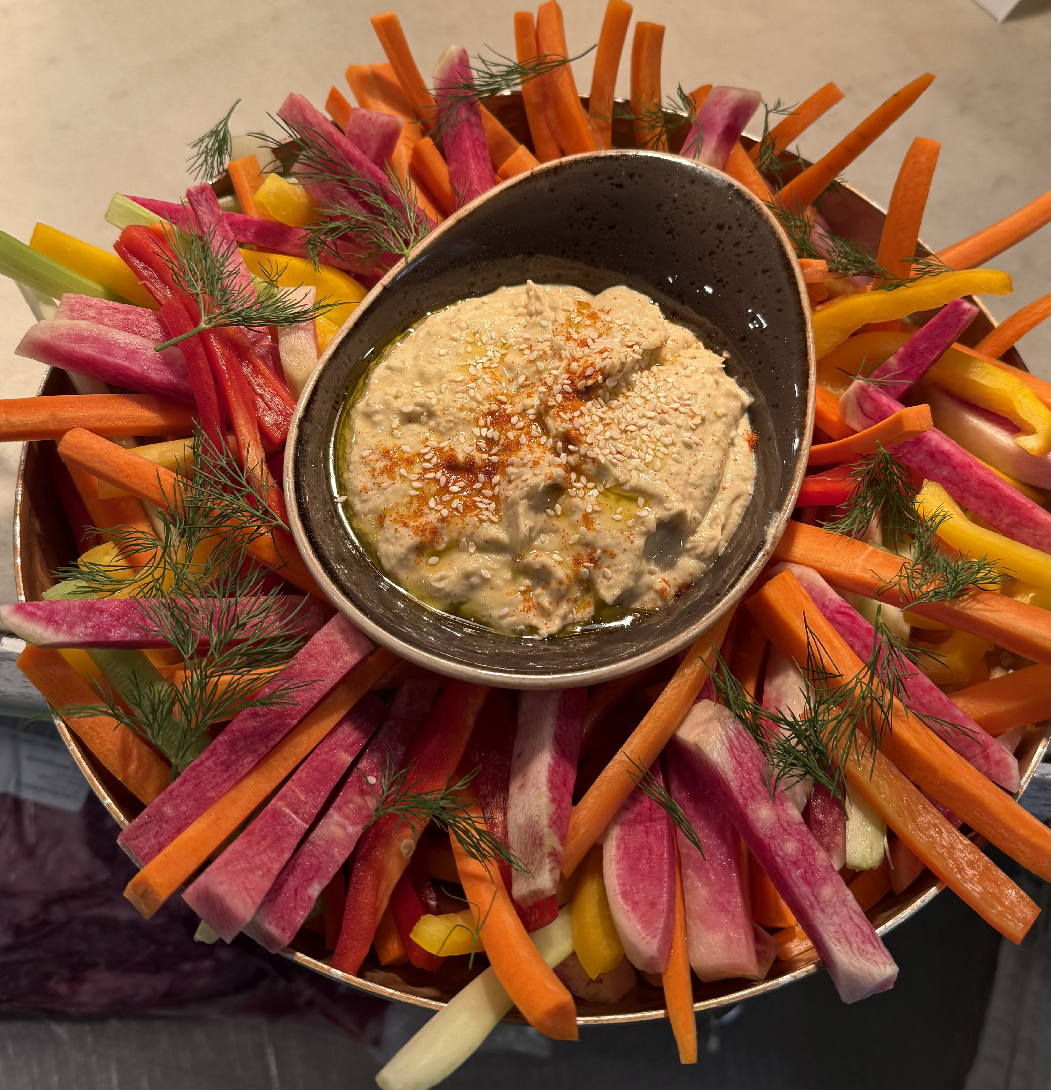 Bowl of creamy hummus topped with sesame seeds, paprika, and olive oil, surrounded by fresh crudités including carrot sticks, radish sticks, celery, and bell pepper slices garnished with dill.