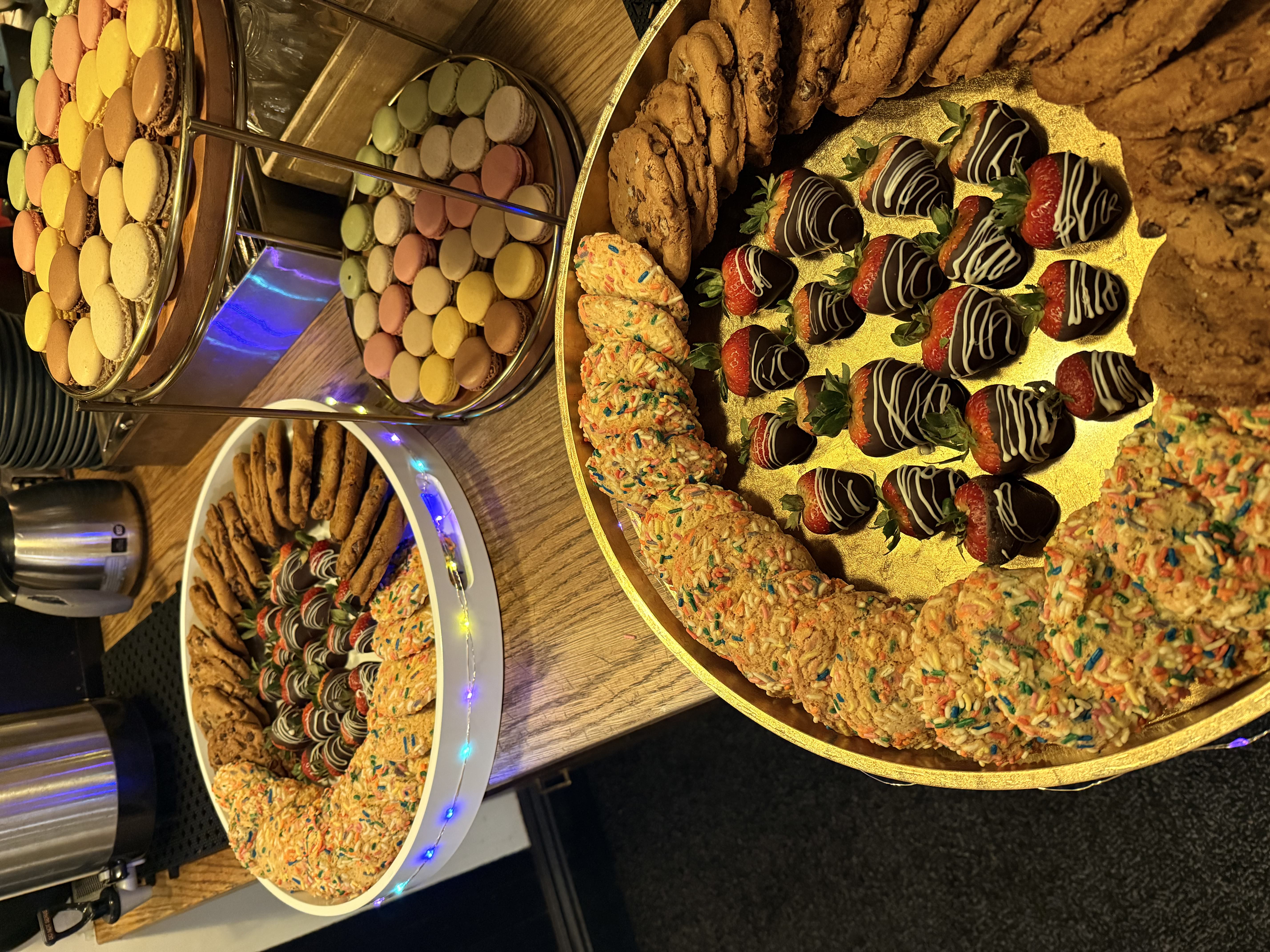 Trays of assorted desserts including chocolate chip cookies, sprinkle cookies, chocolate-covered strawberries, and colorful macarons on a wooden table.