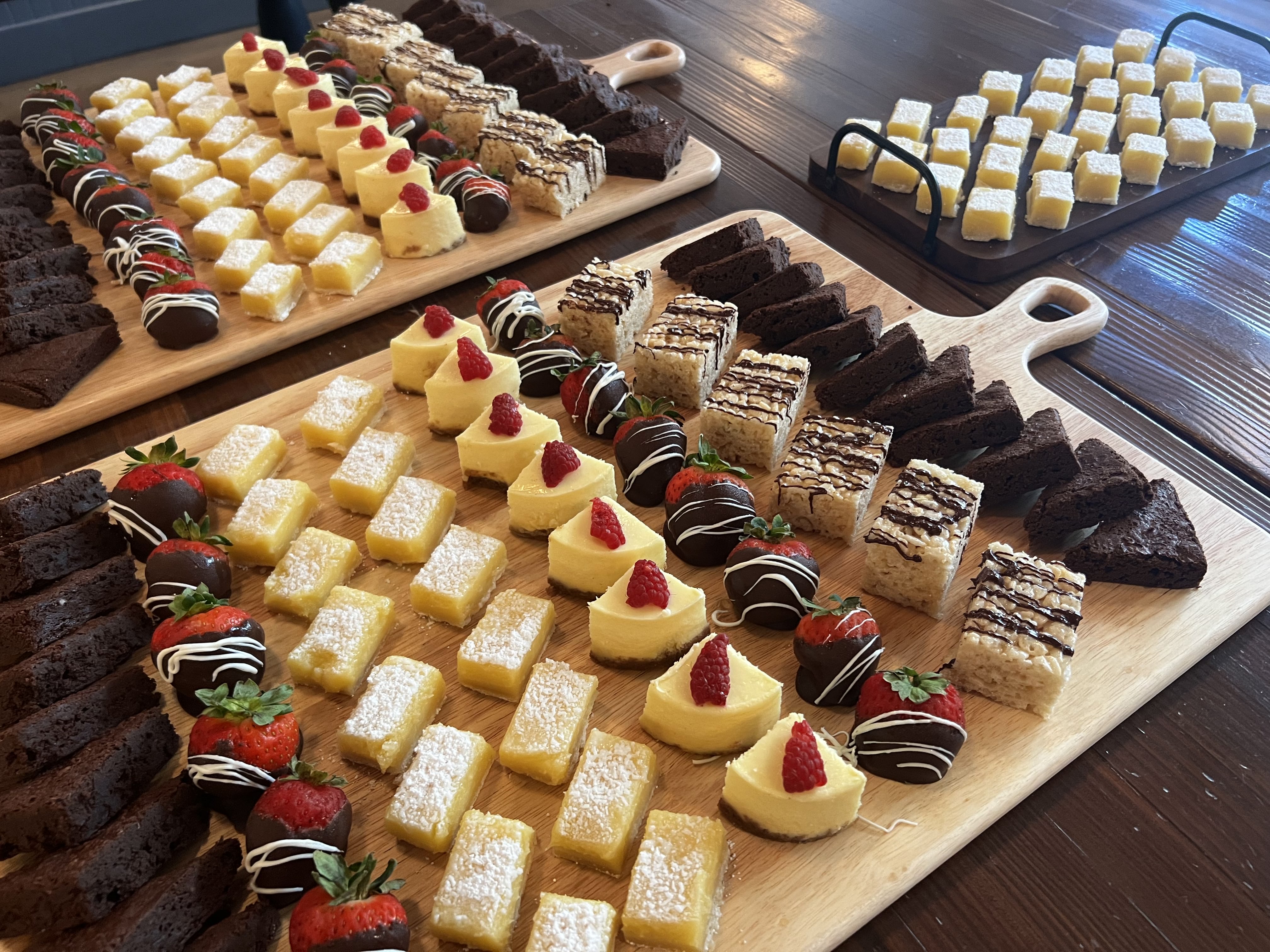 Wooden boards with an assortment of desserts including chocolate brownies, chocolate-covered strawberries, lemon bars dusted with powdered sugar, mini cheesecakes topped with raspberries, and rice crispy treats drizzled with chocolate.