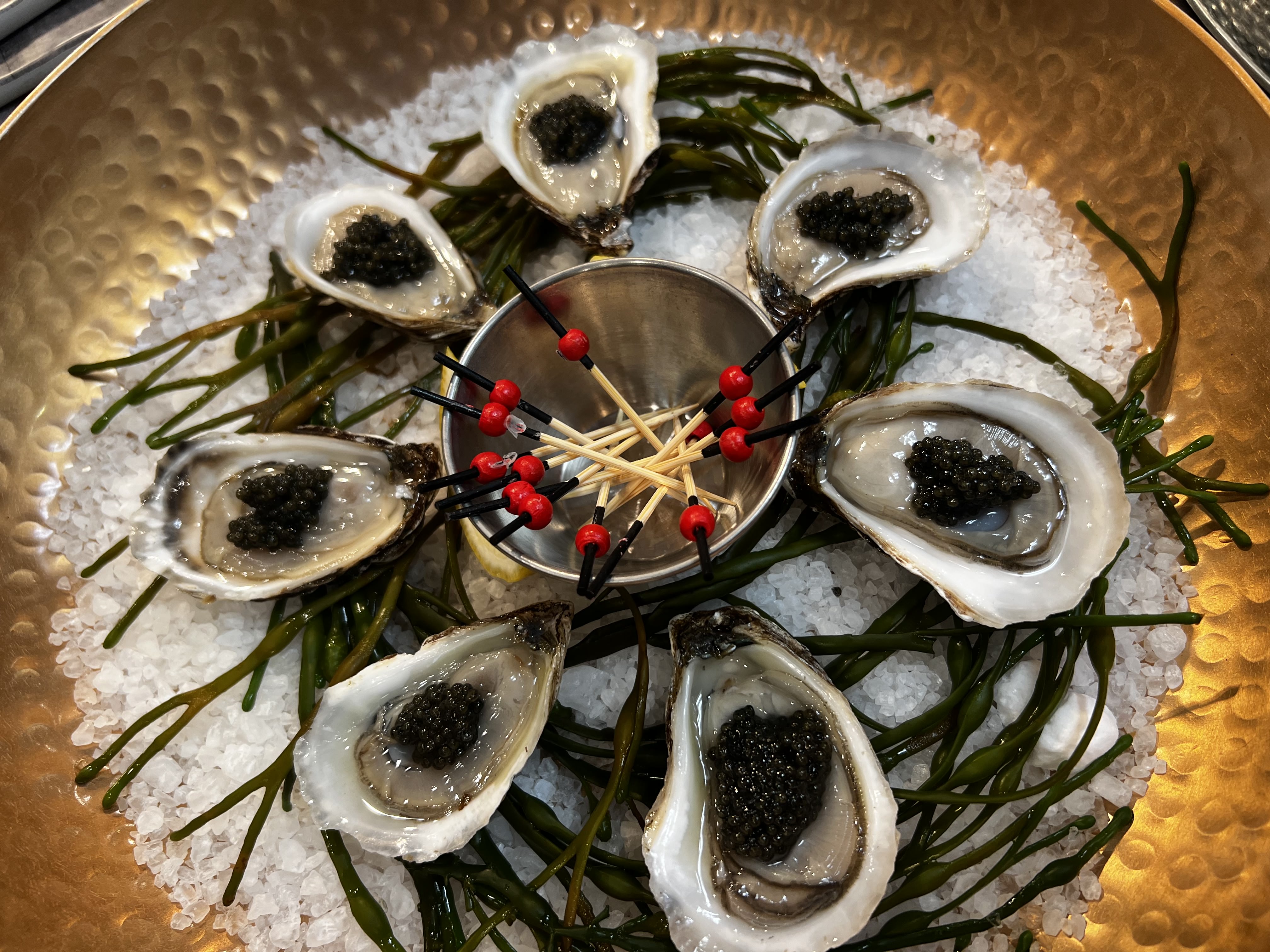Plate of fresh oysters topped with black caviar on a bed of crushed ice and seaweed, with cocktail picks in a small metal bowl at the center.