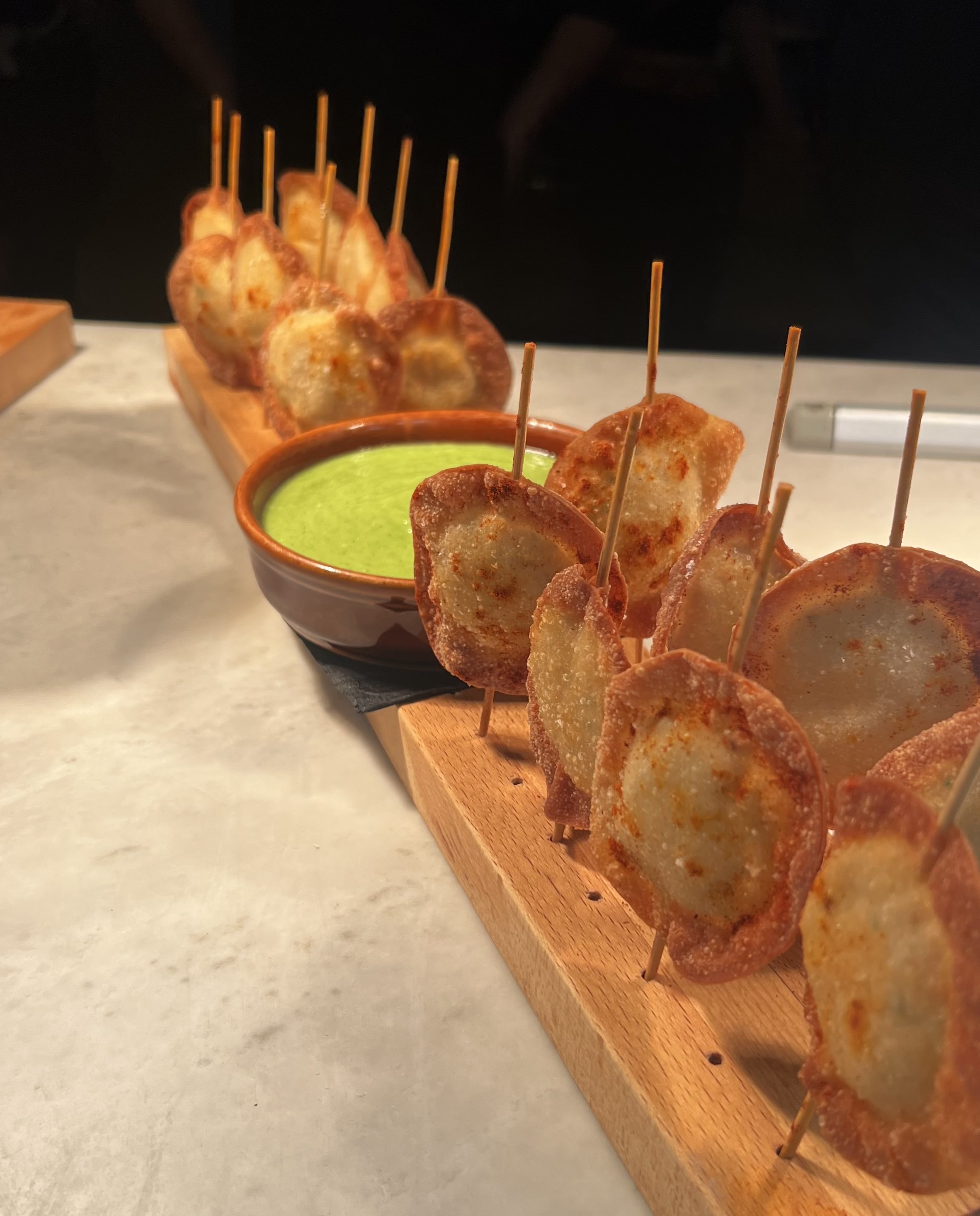 Skewered, golden-brown fried dumplings arranged on a wooden board with a bowl of green dipping sauce.