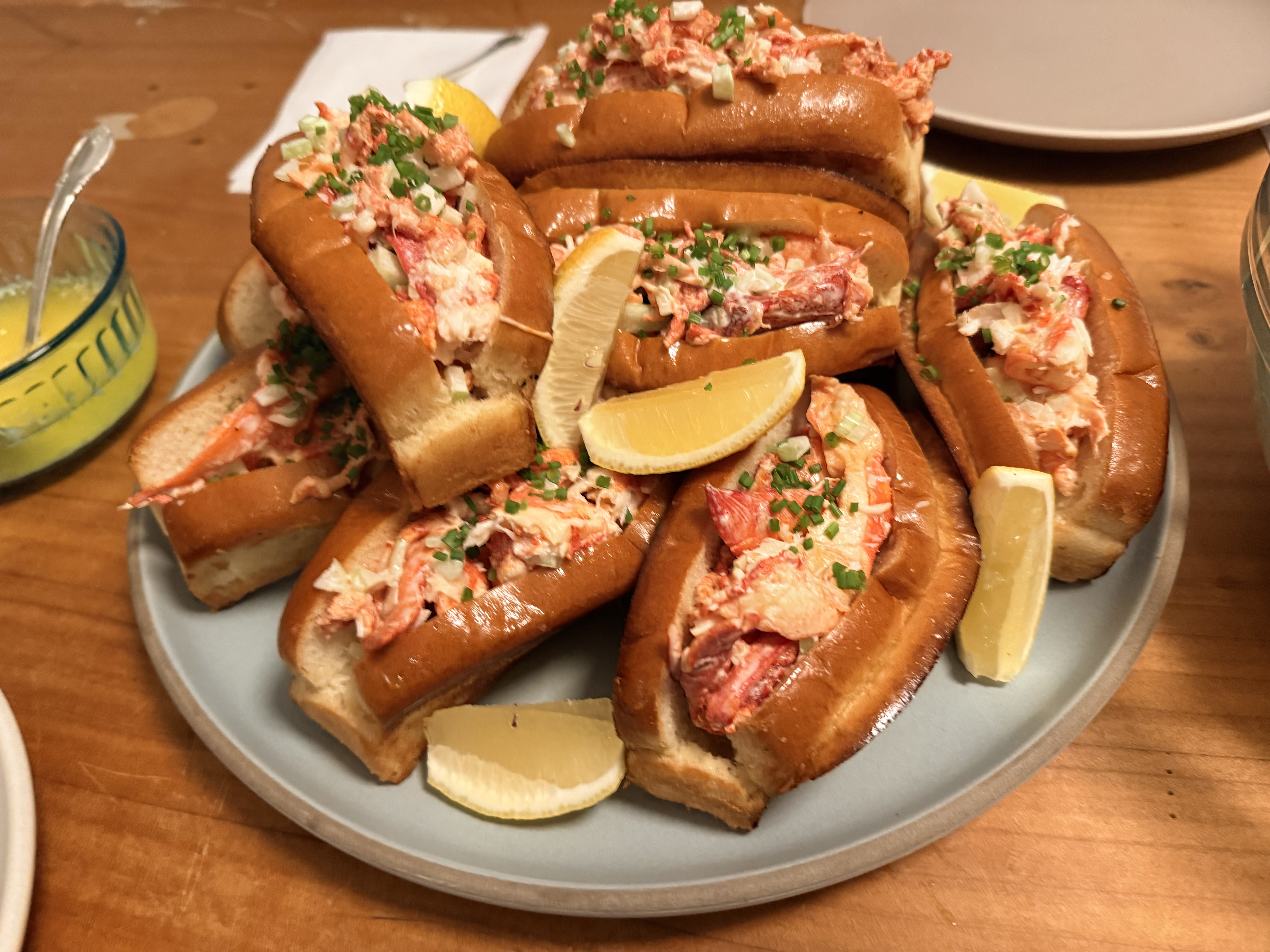 Plate of lobster roll sandwiches garnished with chopped chives and lemon wedges on a wooden table.