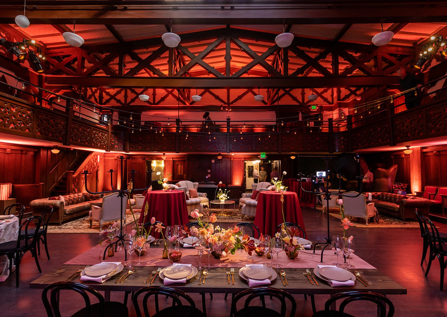 Elegant event space with wooden beams, ambient reddish lighting, round tables with red tablecloths, floral centerpieces, and arranged dining set-up.