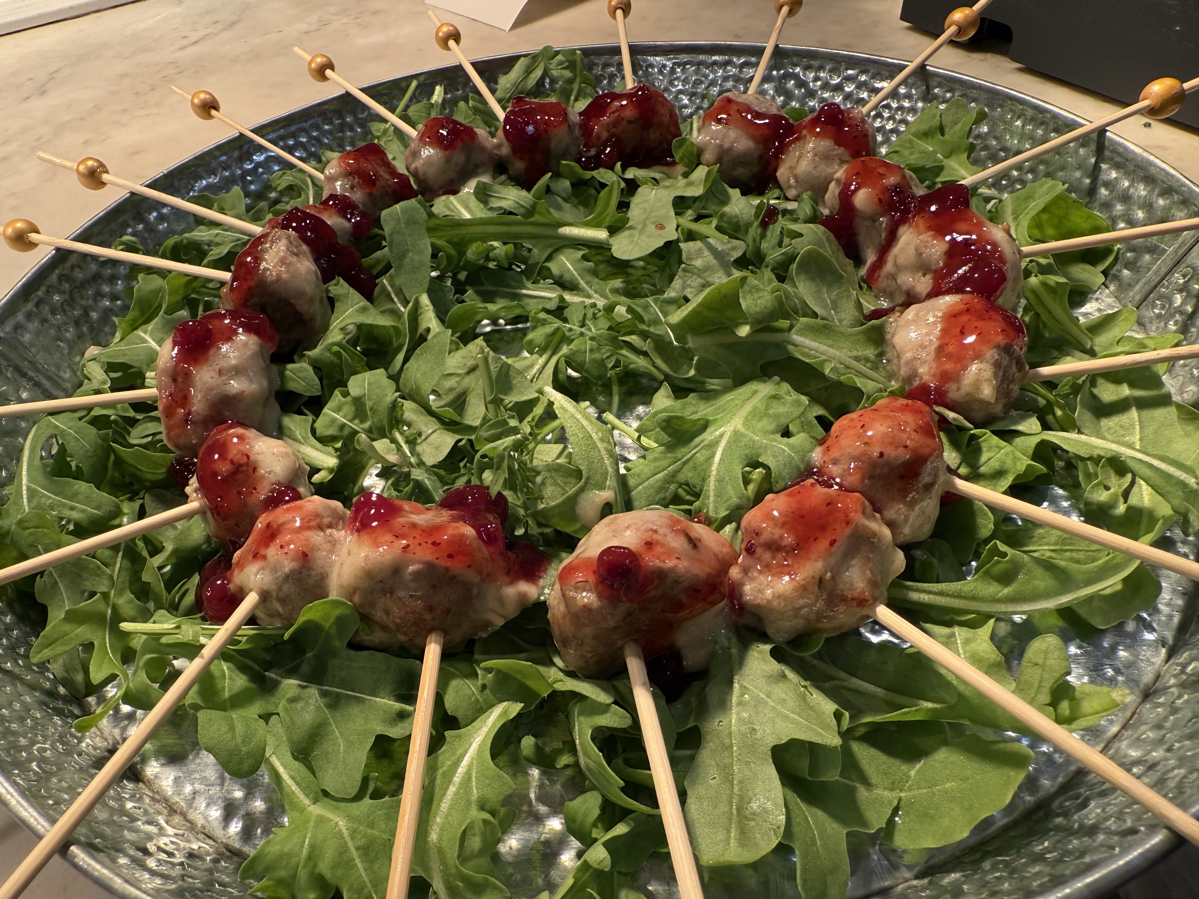 Skewers of meatballs topped with sauce arranged in a circle on a bed of fresh arugula leaves.