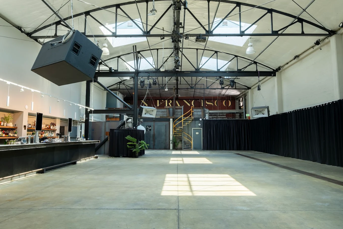Empty industrial-style event space with concrete floor, black curtains, suspended speakers, skylights, and a long bar on the left.