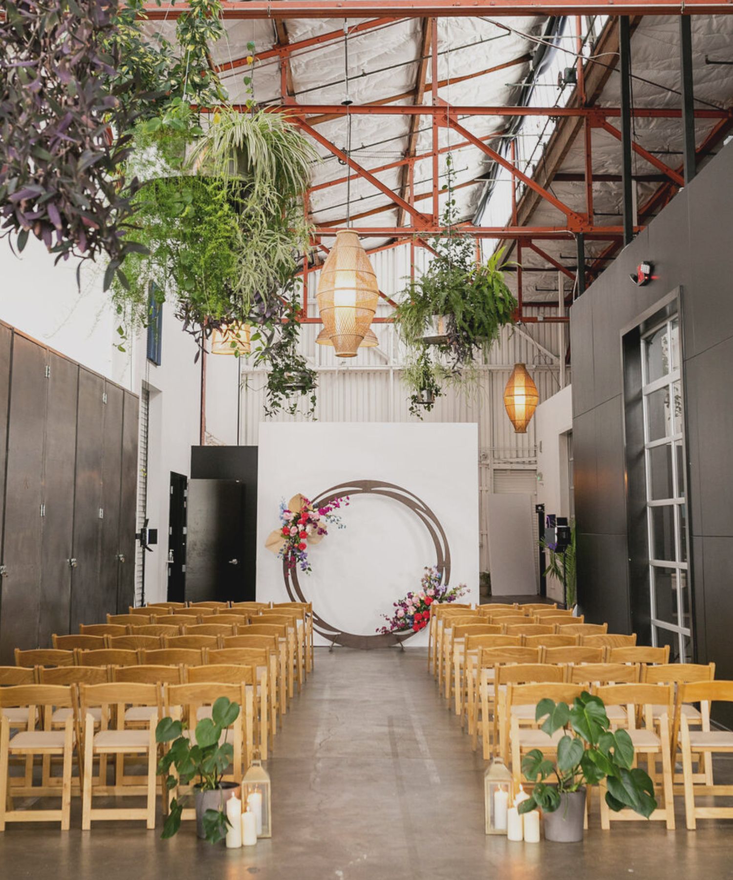 Indoor event space with rows of wooden folding chairs facing a circular floral arch, surrounded by hanging plants and lanterns.