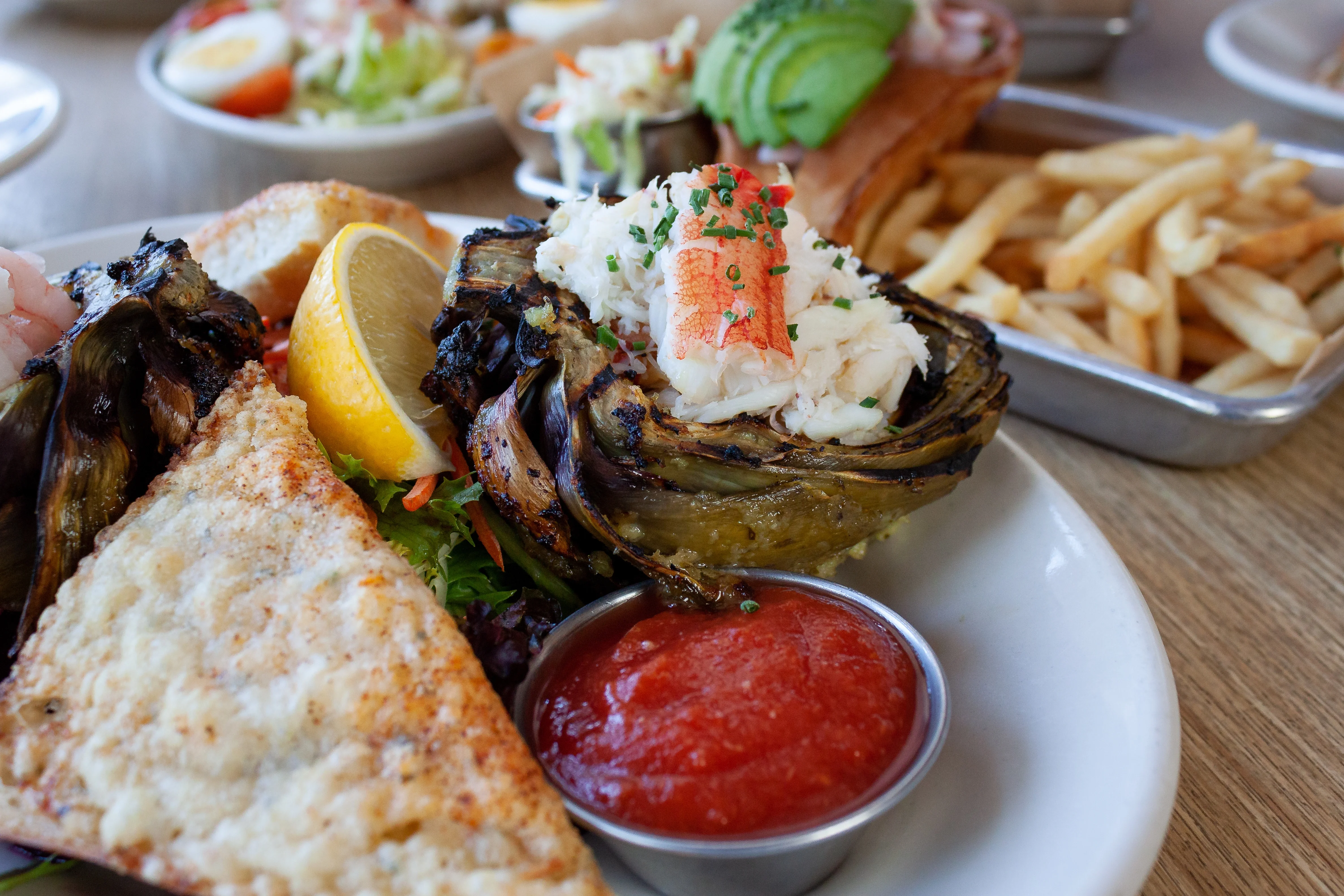 Grilled artichoke topped with crab meat and a lemon wedge on a plate with creamy fish fillet, cocktail sauce, and background tray of fries.