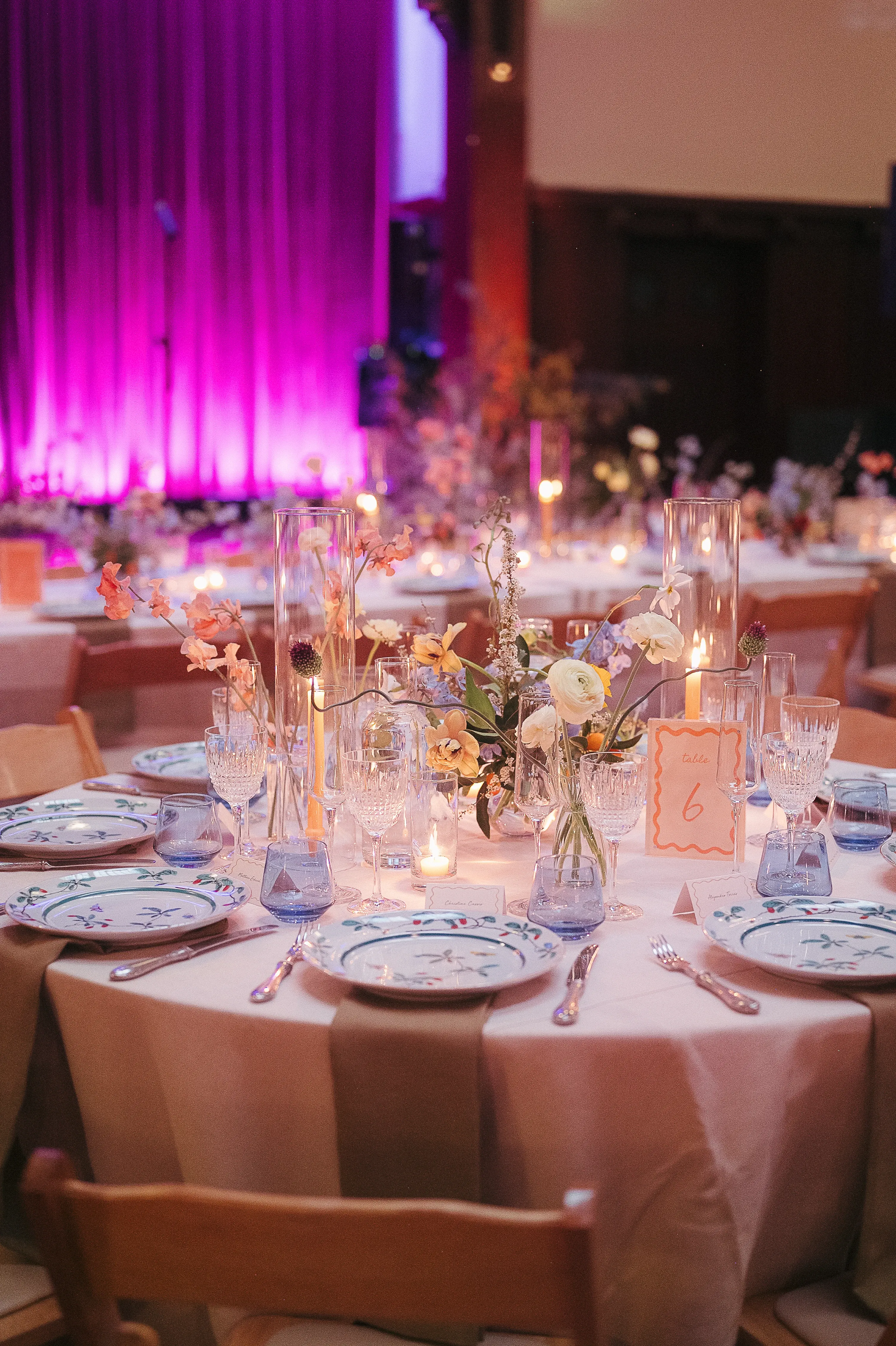 Elegant round table set for a formal event with floral-patterned plates, crystal glasses, candles, and a floral centerpiece under purple lighting.