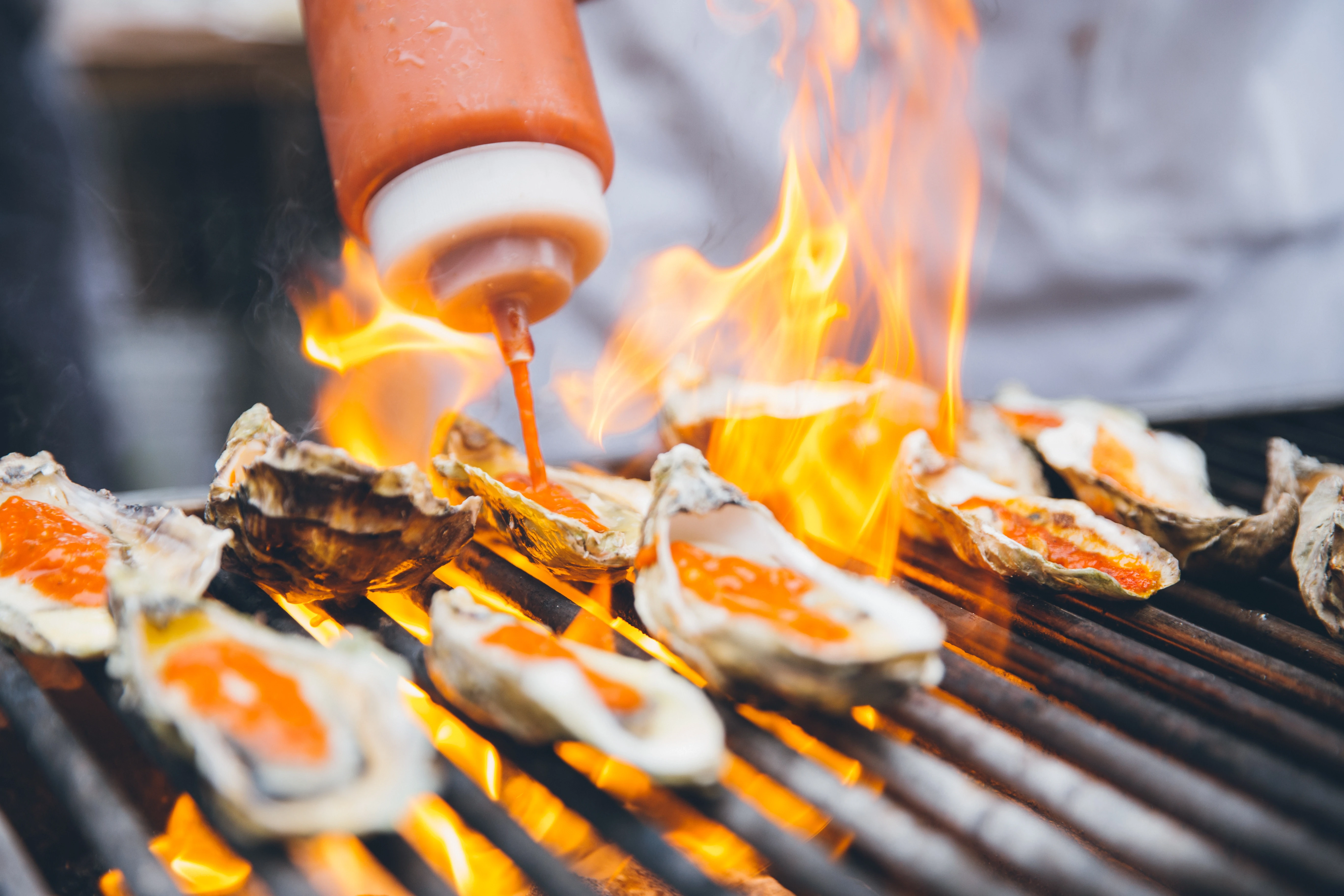 Grilled oysters on a barbecue with orange sauce being squeezed onto them amid flames.