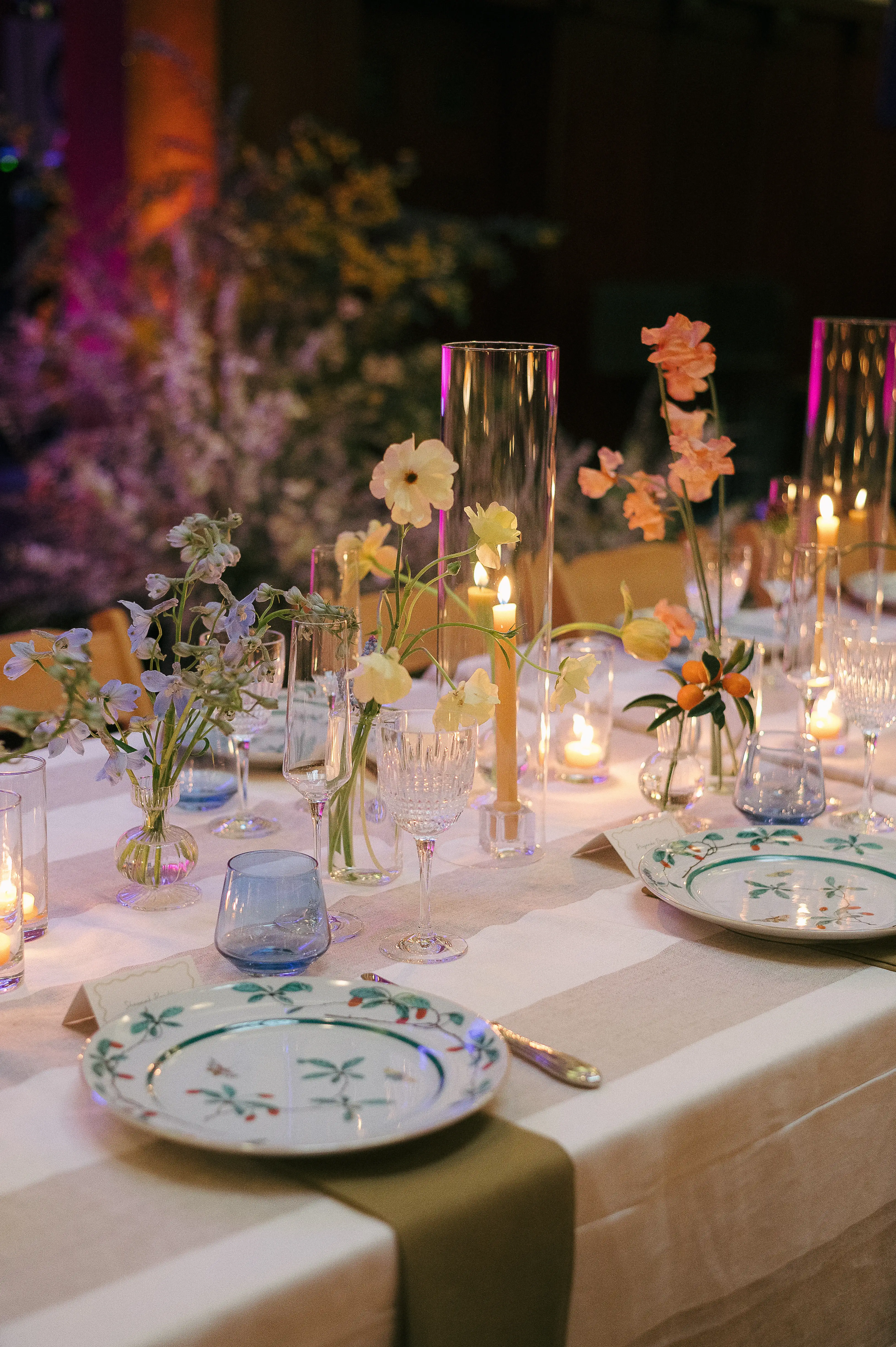 Elegant table setting with floral-patterned plates, glassware, candles, and vases holding delicate flowers on a striped tablecloth.