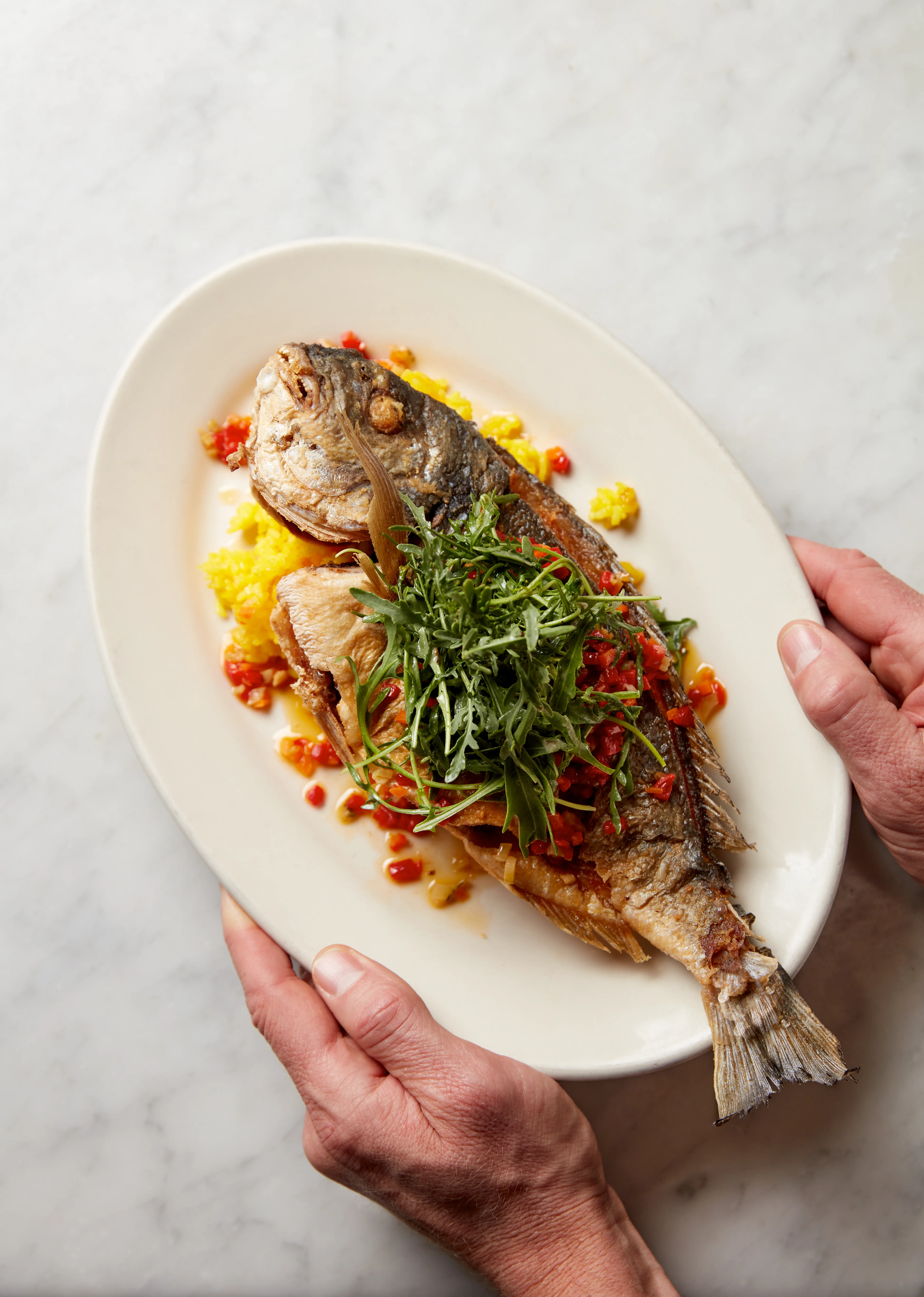 Whole cooked fish served on a white oval plate with yellow rice, chopped red peppers, and topped with fresh arugula, held by two hands.