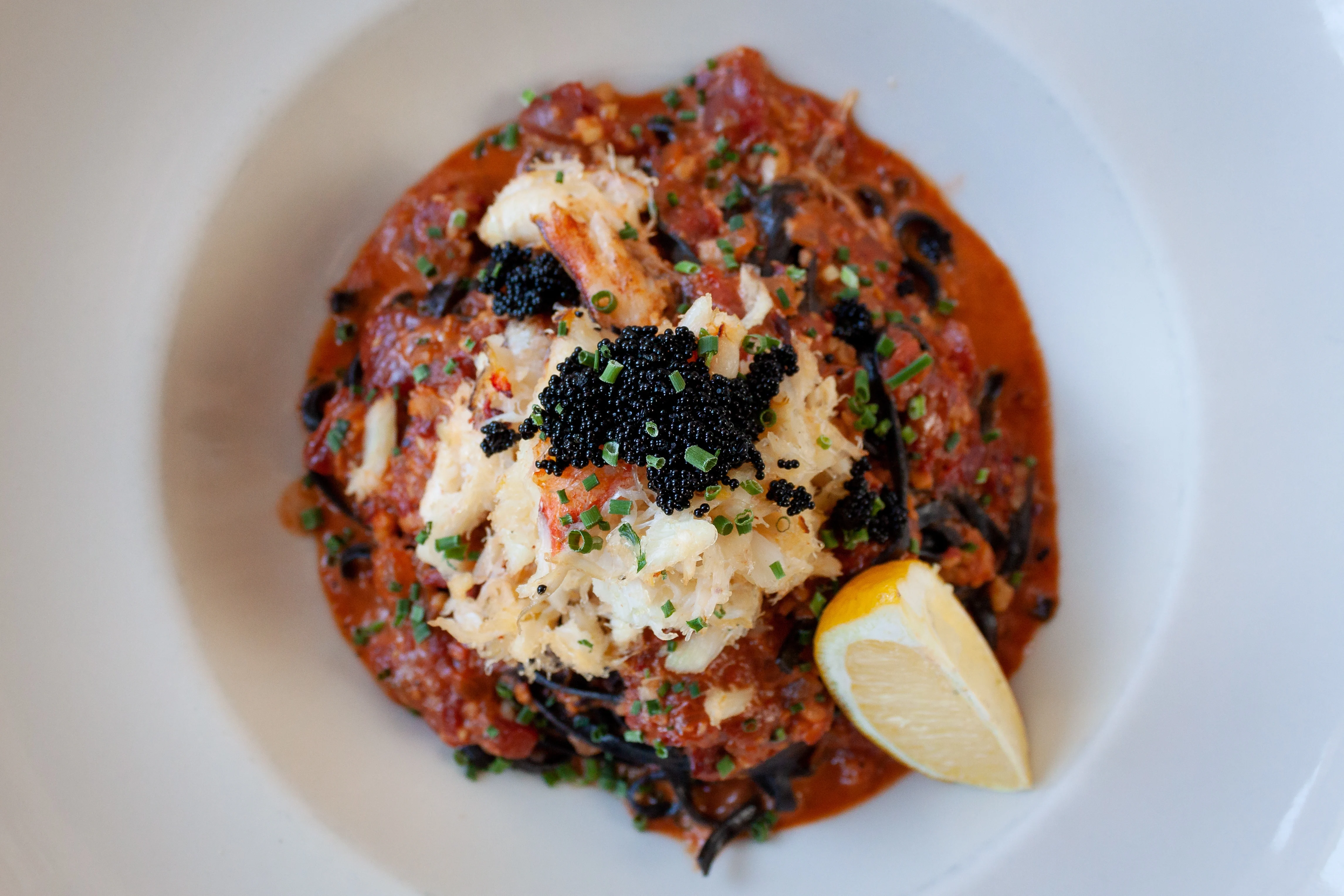 Plate of black pasta topped with crab meat, black caviar, chopped chives, tomato sauce, and a lemon wedge.